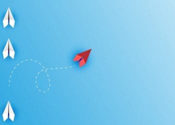 red paper airplane changing course from line of white paper airplanes