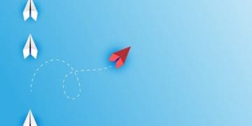 red paper airplane changing course from line of white paper airplanes