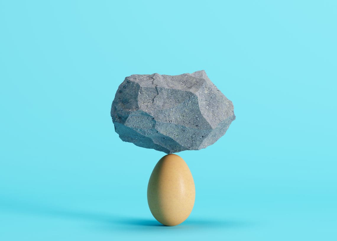 A rock balances on top of an egg in front of a blue background.