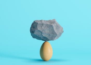 A rock balances on top of an egg in front of a blue background.
