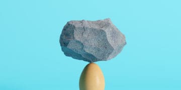 A rock balances on top of an egg in front of a blue background.