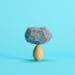 A rock balances on top of an egg in front of a blue background.