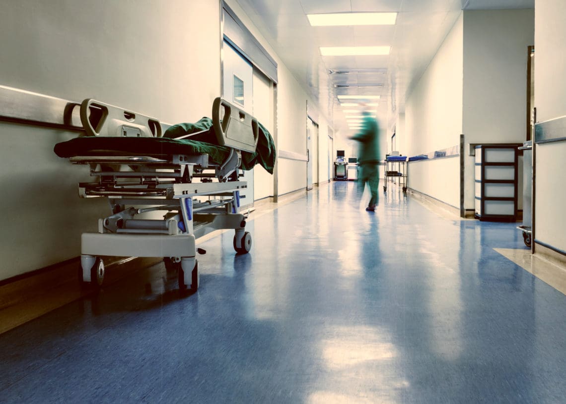 An empty hospital hallway containing a gurney and a doctor in the distance.