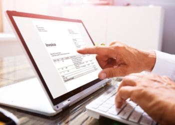 Close-up Of A Businessperson's Hand Analyzing Invoice On Laptop