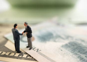 Miniature figures shake hands standing on dollar bills.