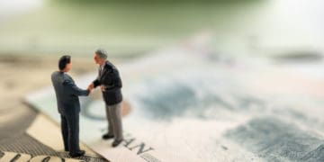 Miniature figures shake hands standing on dollar bills.