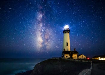 lighthouse shines out at ocean at night under milk way